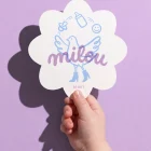 Milou bloemvormig geboortekaartje met een vogel op hoge hakken die babyspulletjes jongleert in lichtblauw, en de naam in lila handschrift met overdruk